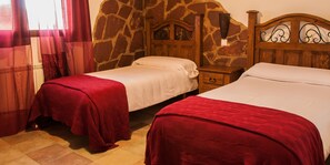 A room with two beds, each with a red blanket, a stone wall, a wooden headboard, and a nightstand with a lamp. - Casa Rural La Dueña (El Pobo de Duenas)