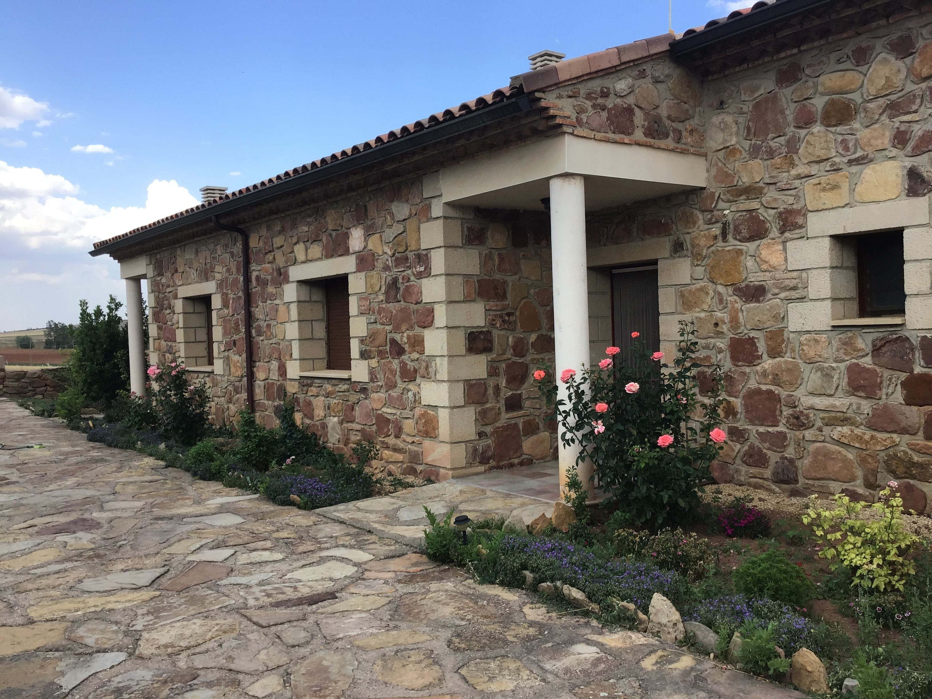 Un edificio in pietra con tetto a tegole, un vialetto in pietra e un giardino con diverse piante e fiori.