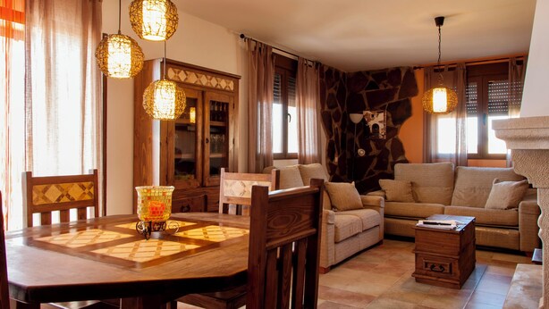 A cozy living room with a wooden dining table, a sofa, and a coffee table. There are hanging lights and a fireplace. - Casa Rural La Dueña (El Pobo de Duenas)