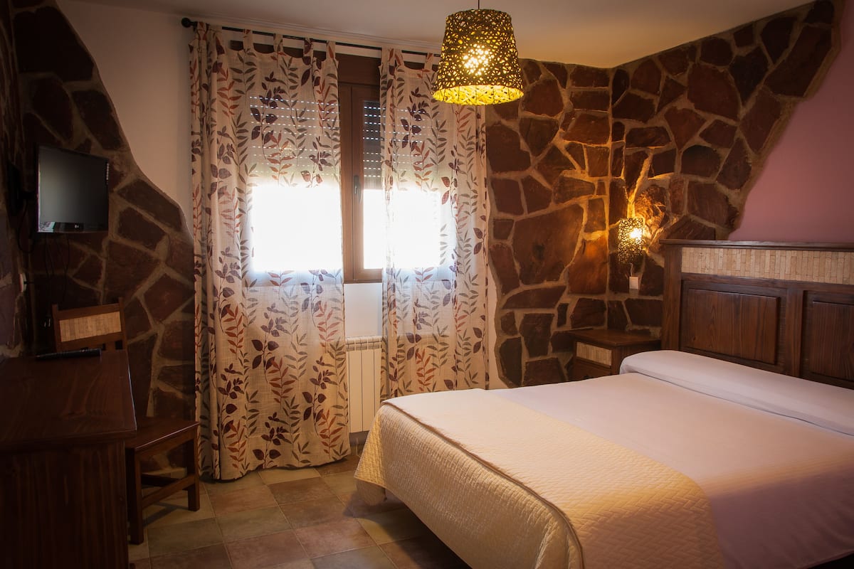 A bedroom with a stone wall, a bed, a television, and a window with floral curtains.