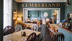 En matplats med en skylt som visar texten ”CUMBERLAND”, träbord, stolar, en skylt med texten ”VÄLKOMMEN”, en klocka och en bardisk med flaskor och glas.