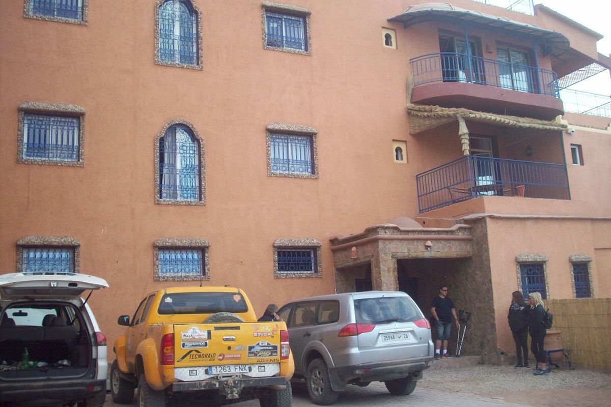 A multi-story building with a red facade and multiple windows. There are several vehicles parked in front, including a yellow pickup truck and a silver SUV.