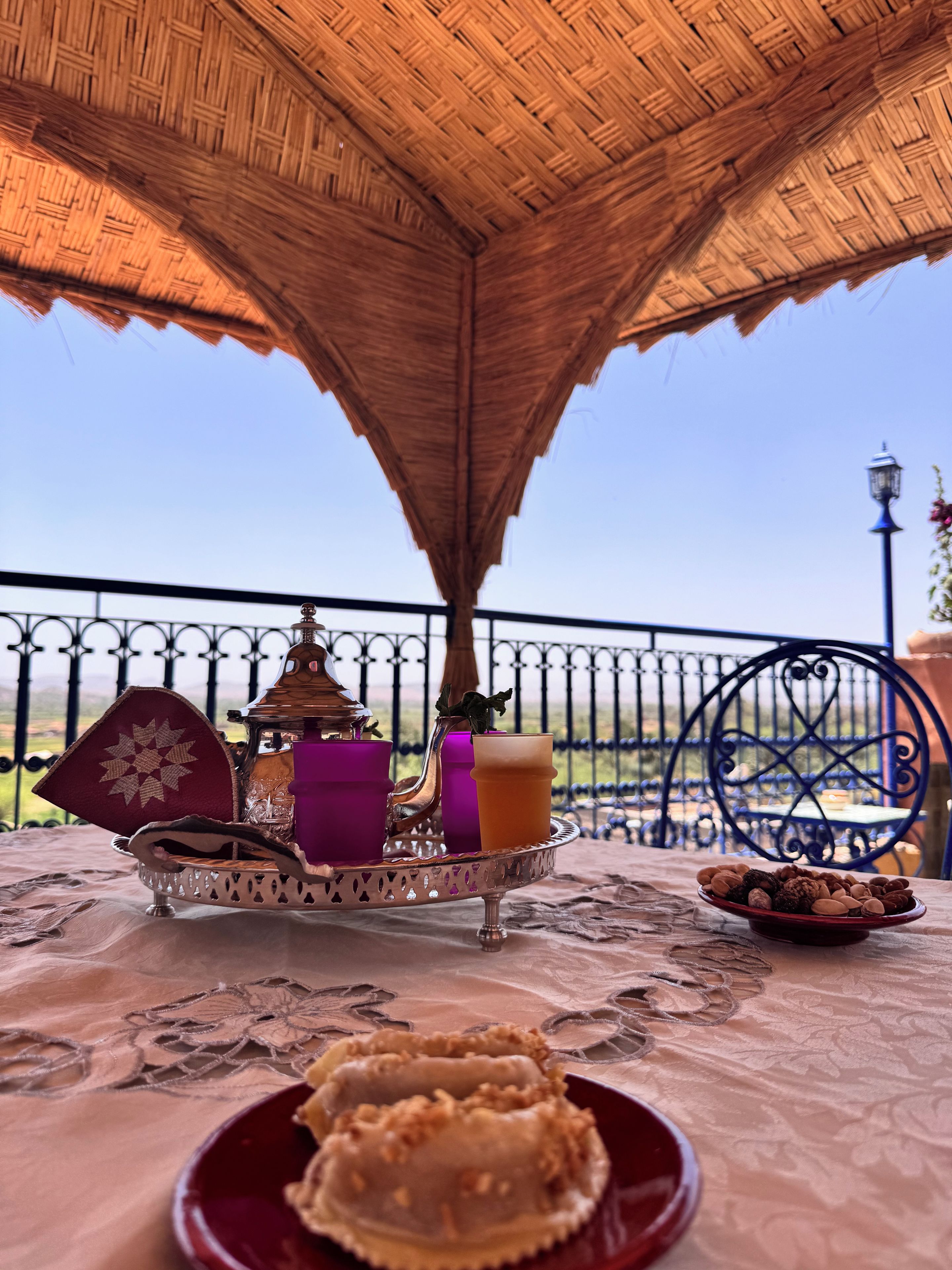 A table with a decorative tray, a candle, a glass, and a plate of pastries under a thatched roof.