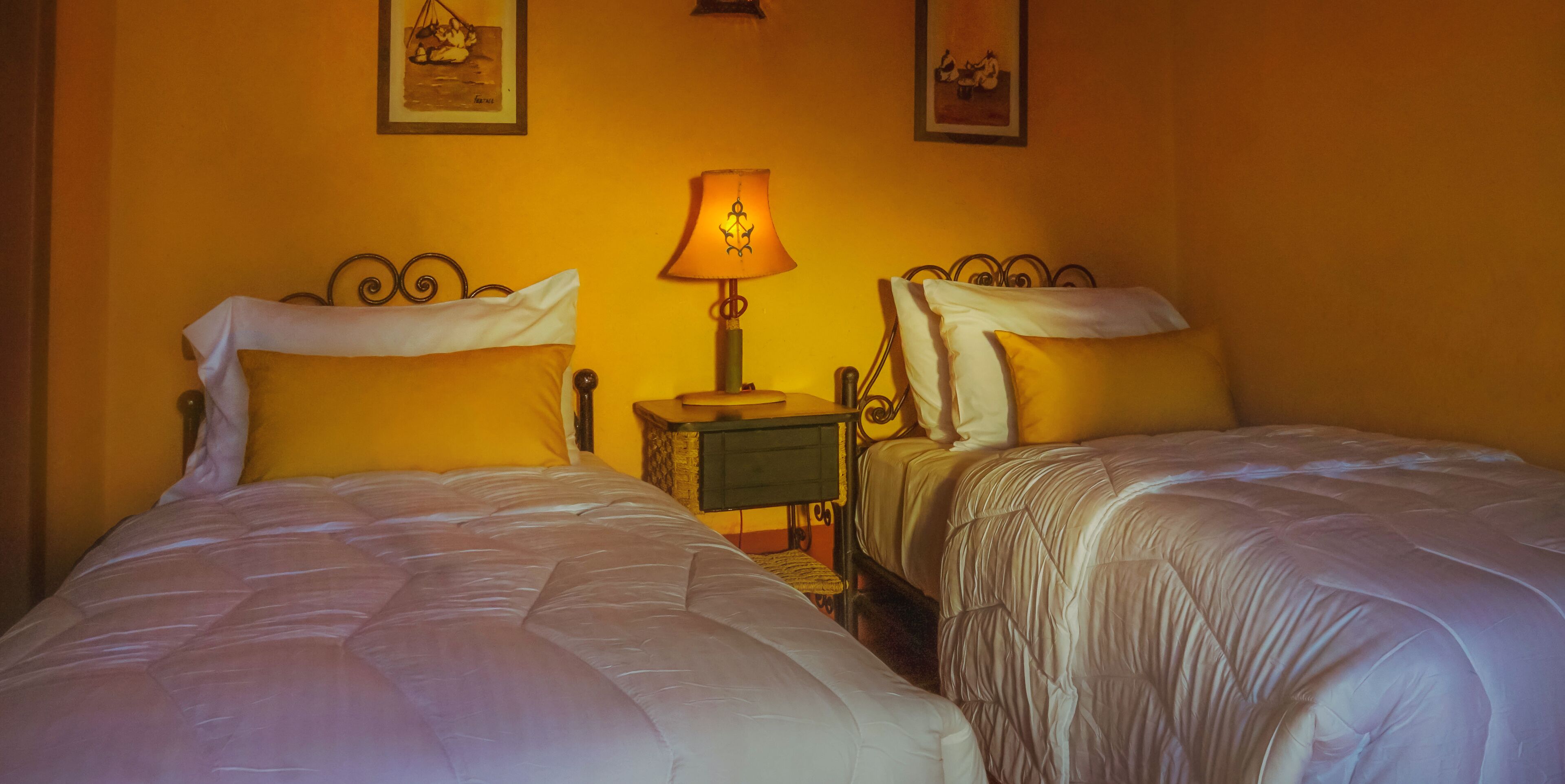 A hotel room with two beds, a nightstand with a lamp, and two framed pictures on the wall.