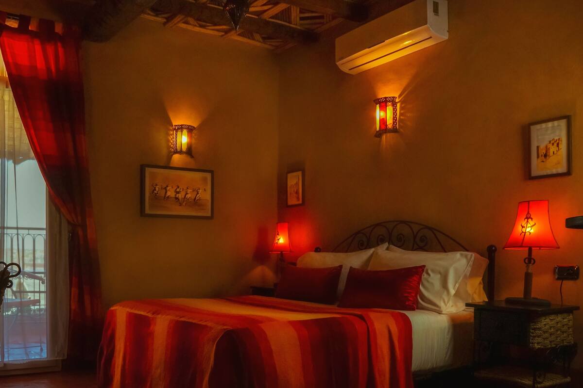 A bedroom with a bed, two wall-mounted lamps, a window with red curtains, and a balcony with a chair.