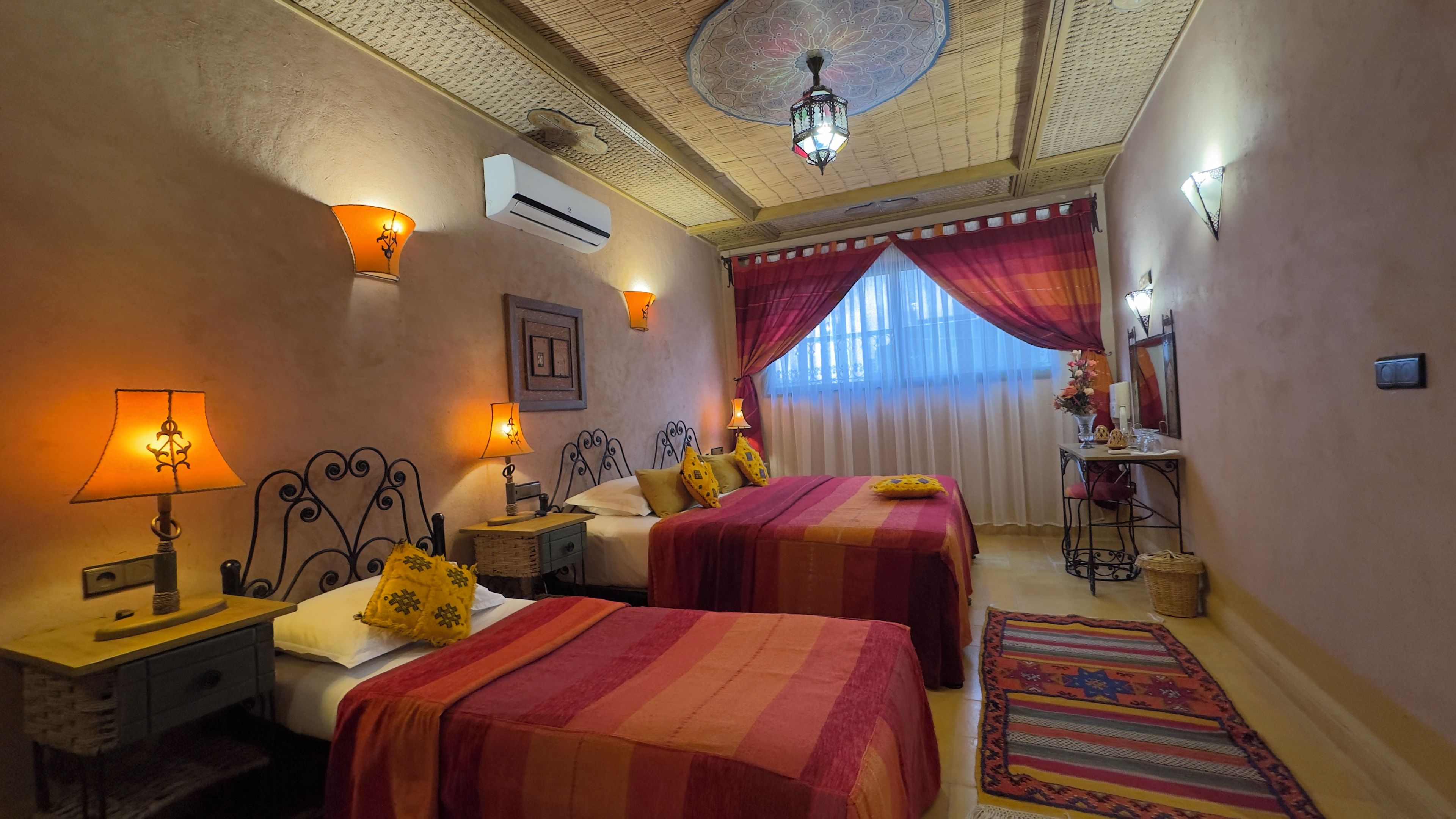 A hotel room with two beds, a desk, a chair, a lamp, an air conditioner, and a patterned rug.