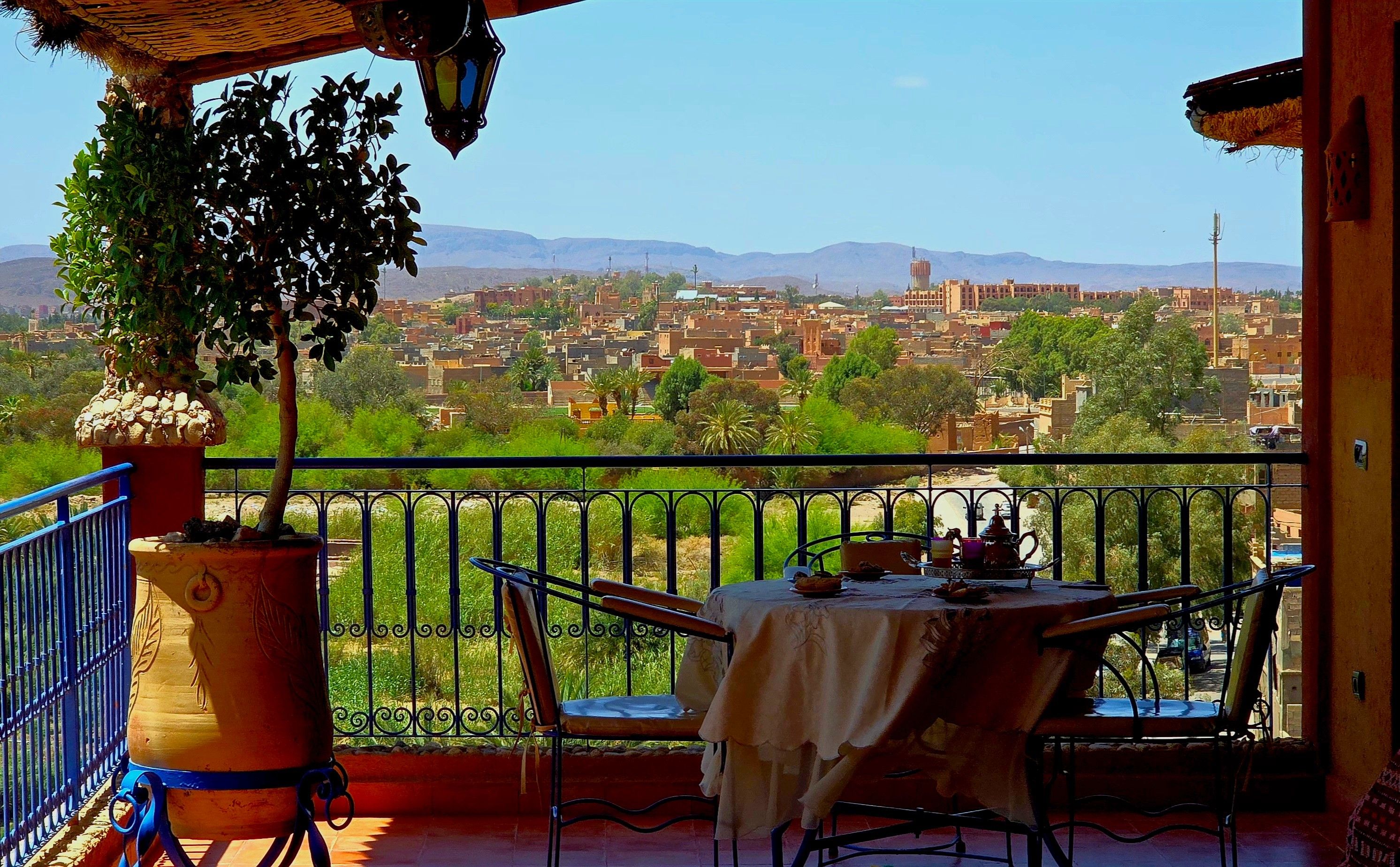 A balcony with a table and chairs, overlooking a cityscape with buildings and mountains.