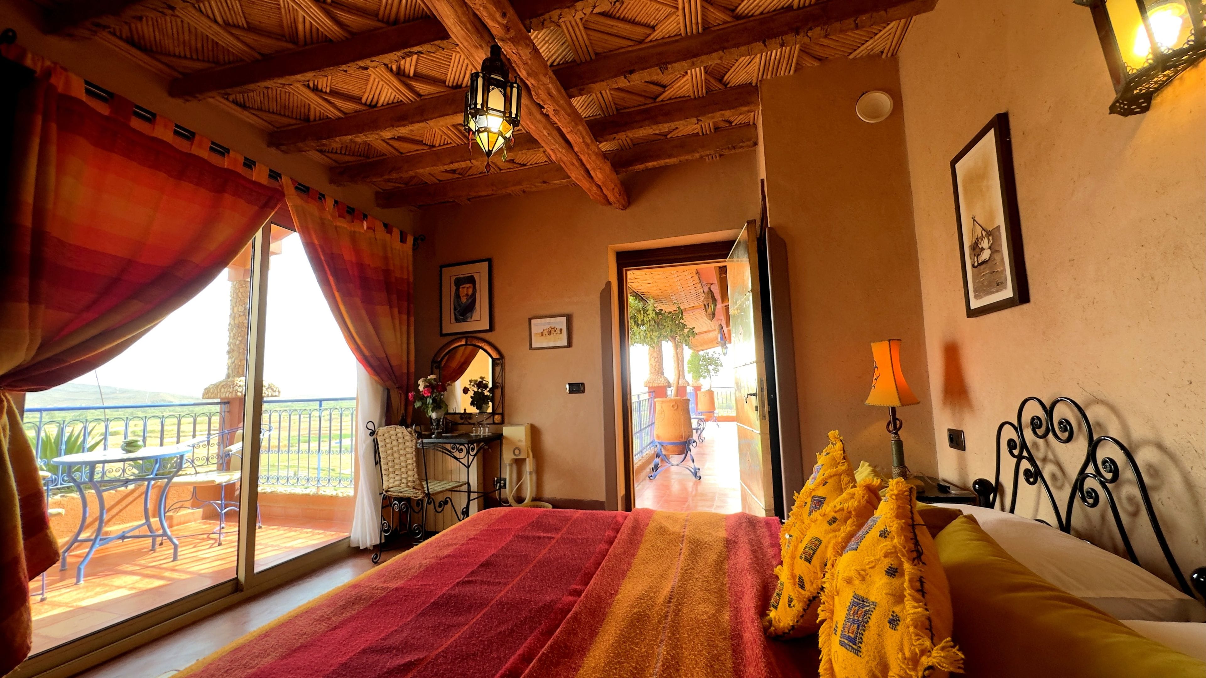 A bedroom with a large bed, a balcony with a table and chairs, and a view of the outdoors.