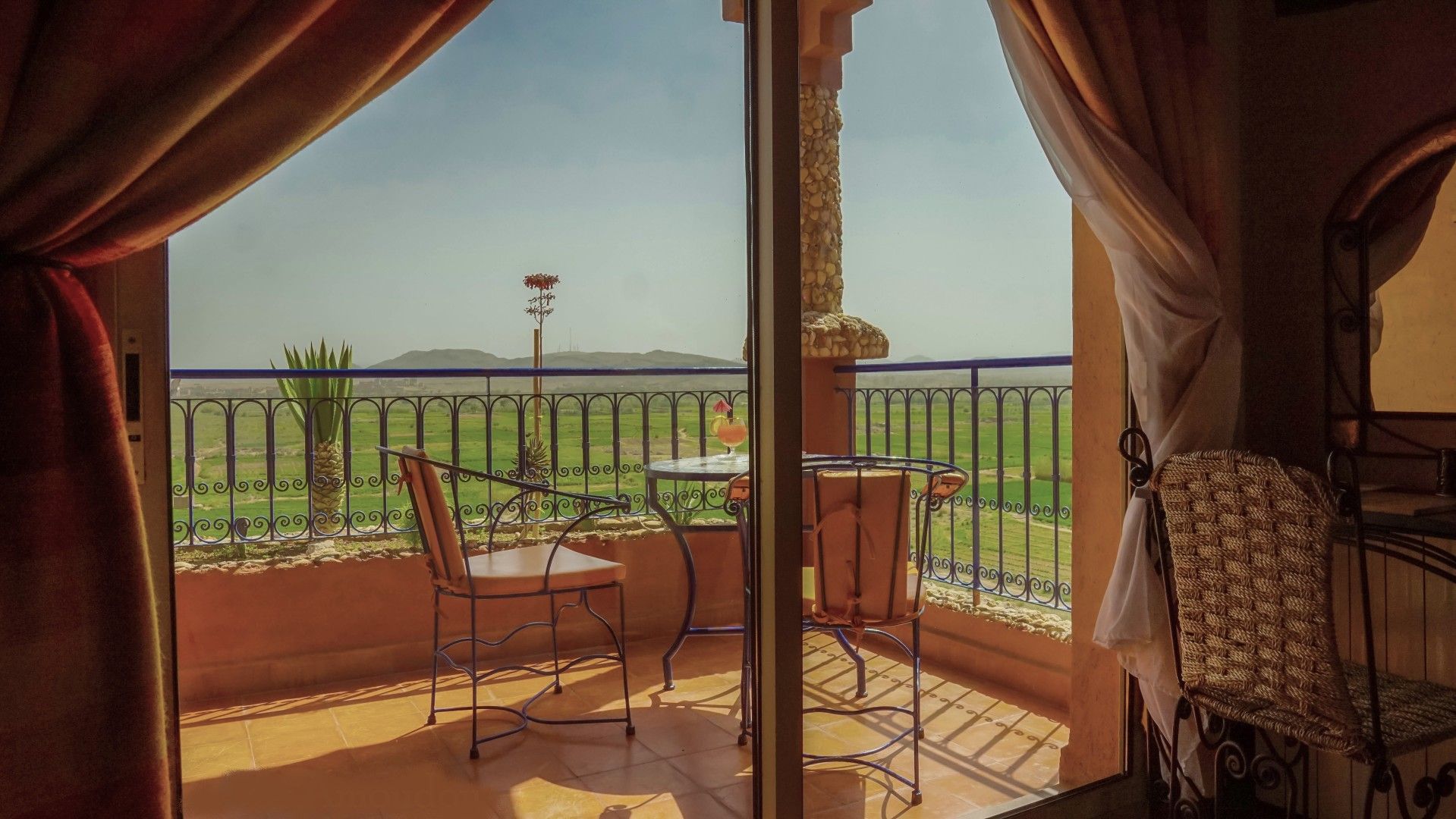 A balcony with a table and chairs, a view of a green landscape, and a decorative plant.