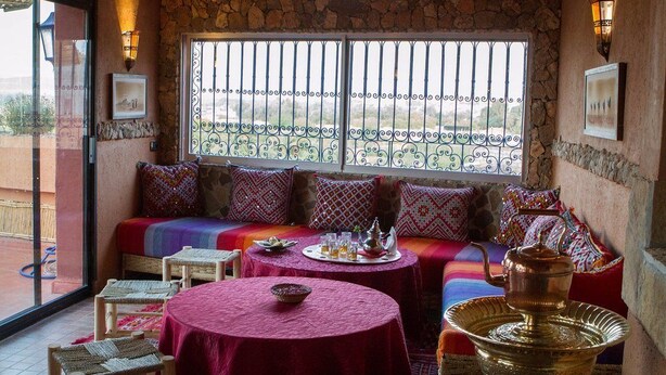 Una habitación acogedora con pared de piedra, un ventanal con celosías de hierro decorativas, un sofá con cojines de colores, una mesa redonda con una bandeja y una tetera de bronce tradicional.