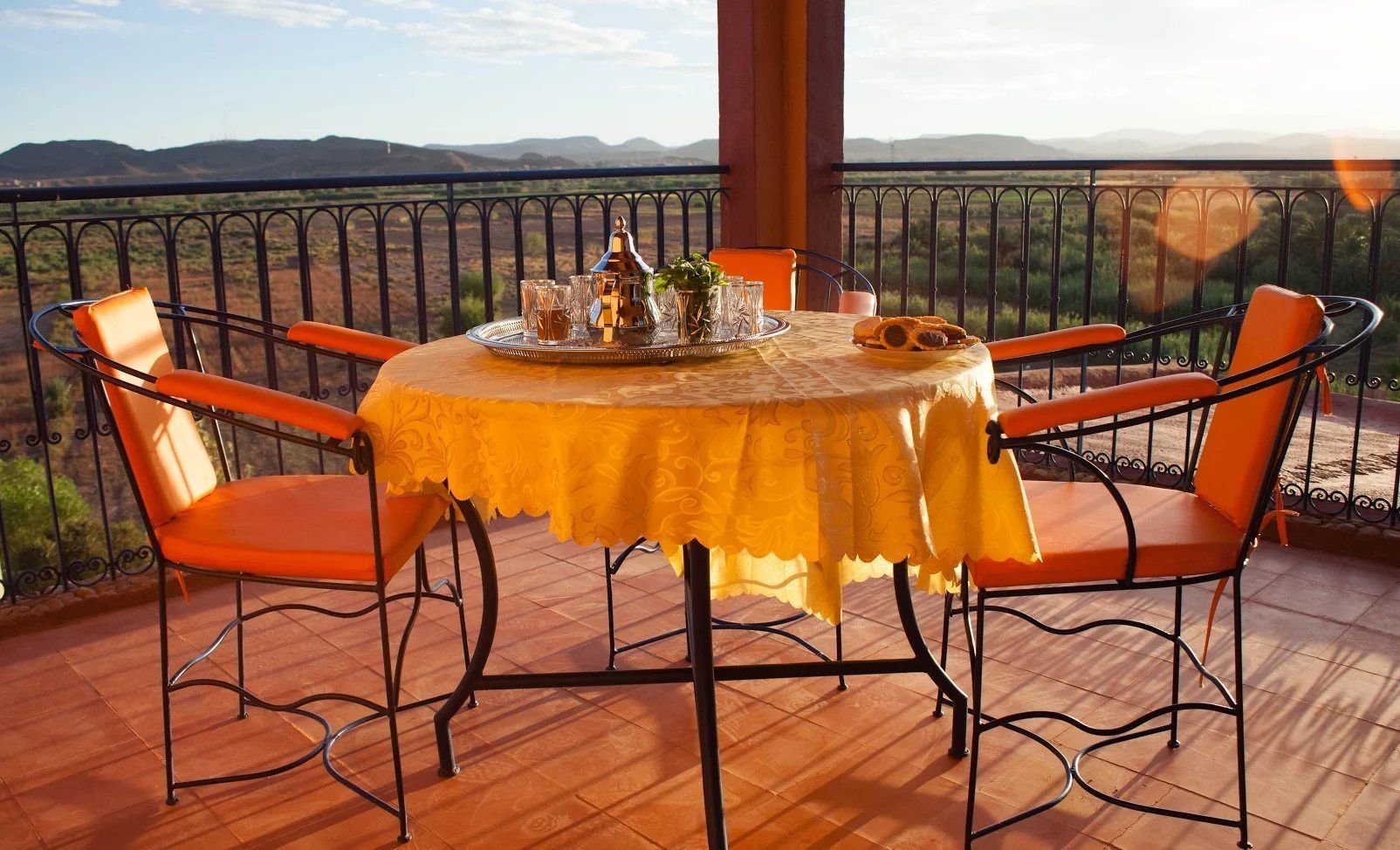 Mesa redonda con mantel amarillo, rodeada por cuatro sillas naranjas de estructura negra, ubicada en un balcón con vistas a colinas distantes.