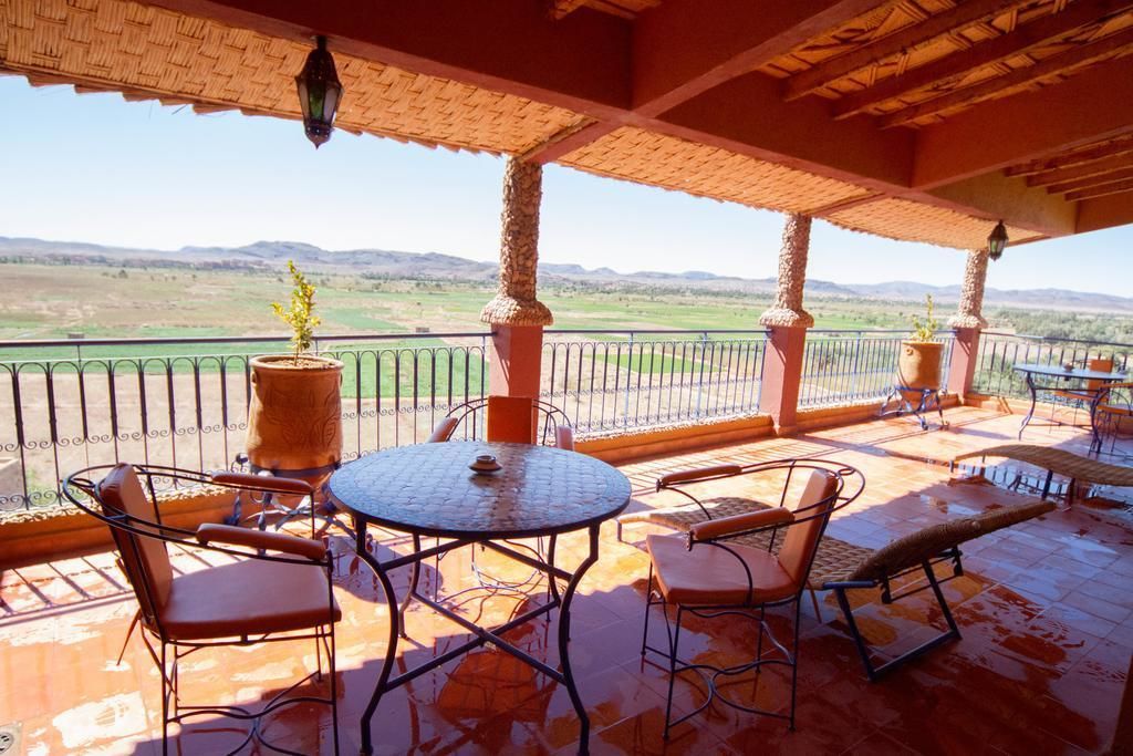A covered outdoor patio with a round table and chairs, potted plants, and a view of the countryside.