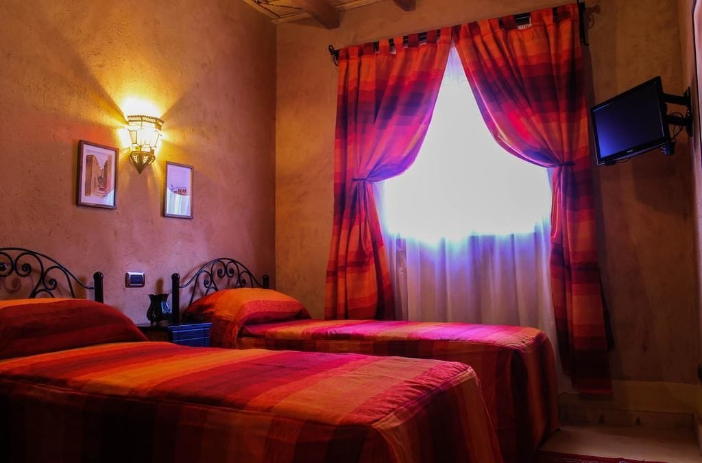Una habitación con dos camas, un televisor en la pared y una ventana con cortinas rojas.