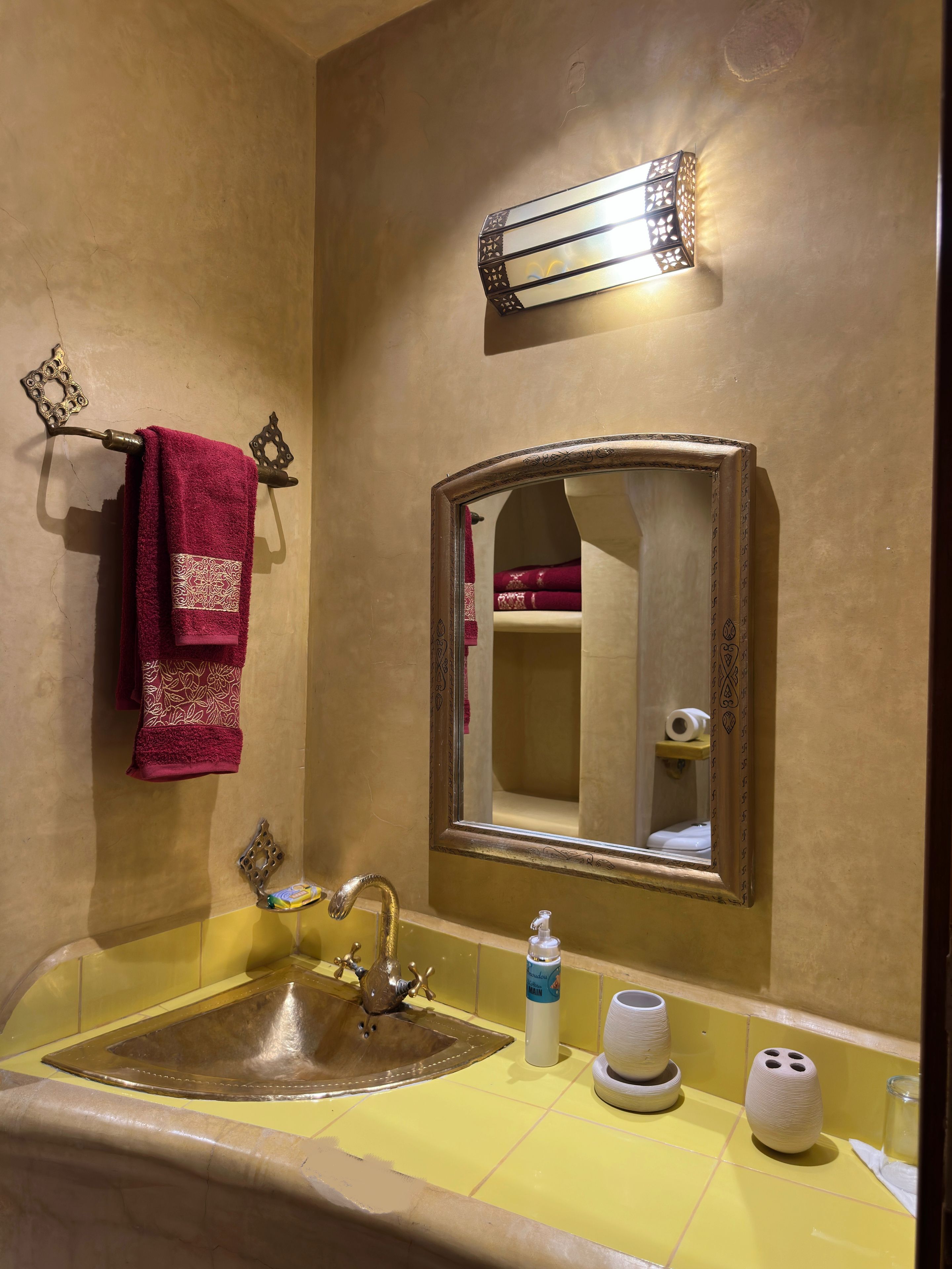 A bathroom with a gold-colored sink, a mirror with an ornate frame, and a towel rack with a red towel.
