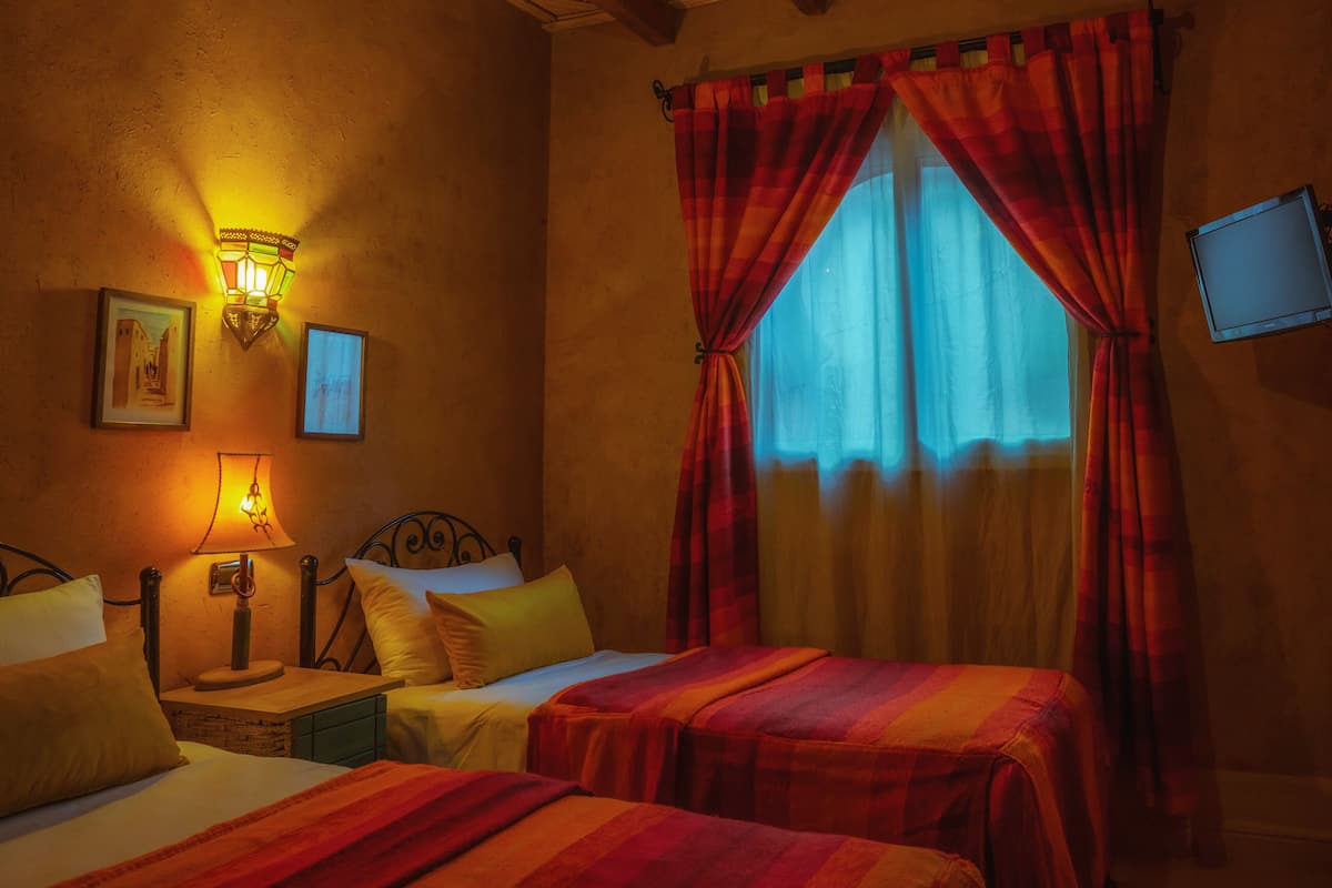 A hotel room with two beds, a nightstand, a lamp, a wall-mounted TV, and a window with red curtains.