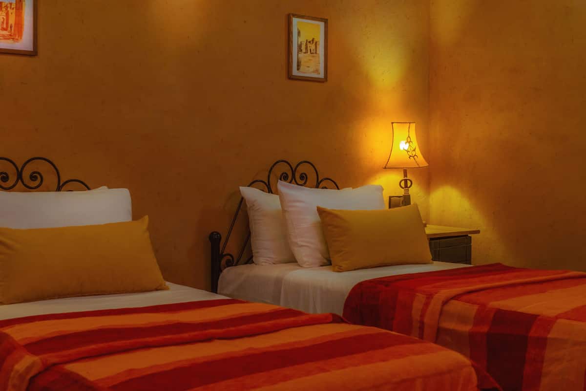 A hotel room with two beds, each with white linens and yellow pillows. There is a nightstand with a lamp between the beds.
