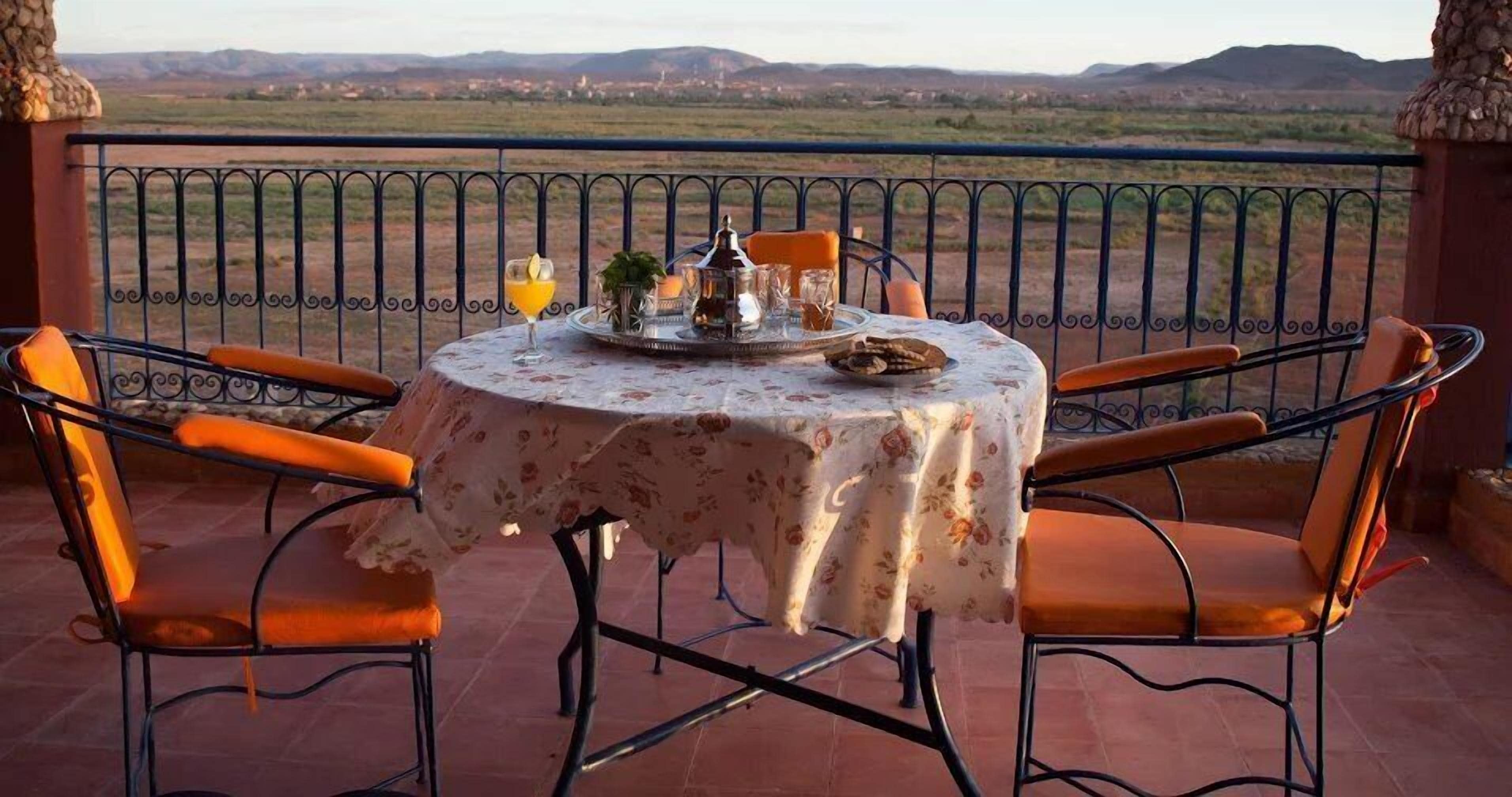 Une table ronde recouverte d’une nappe à motifs floraux, deux chaises orange et une vue sur un paysage montagneux.