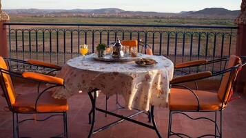 Une table ronde recouverte d’une nappe à motifs floraux, deux chaises orange et une vue sur un paysage montagneux.