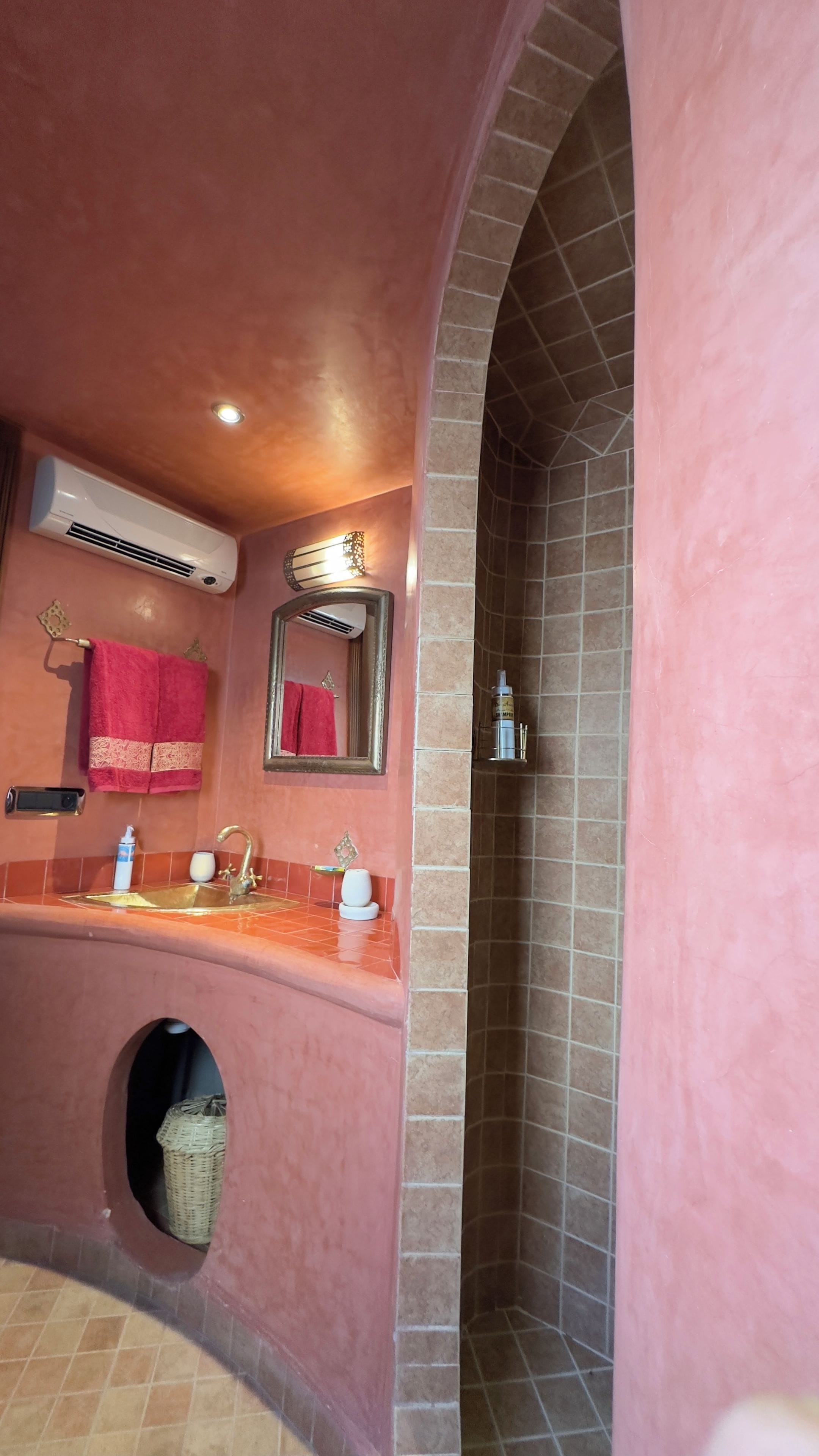 Un baño con pared rosa, un lavabo con grifo dorado, un espejo, un perchero con una toalla roja y una zona de ducha con paredes de azulejos.