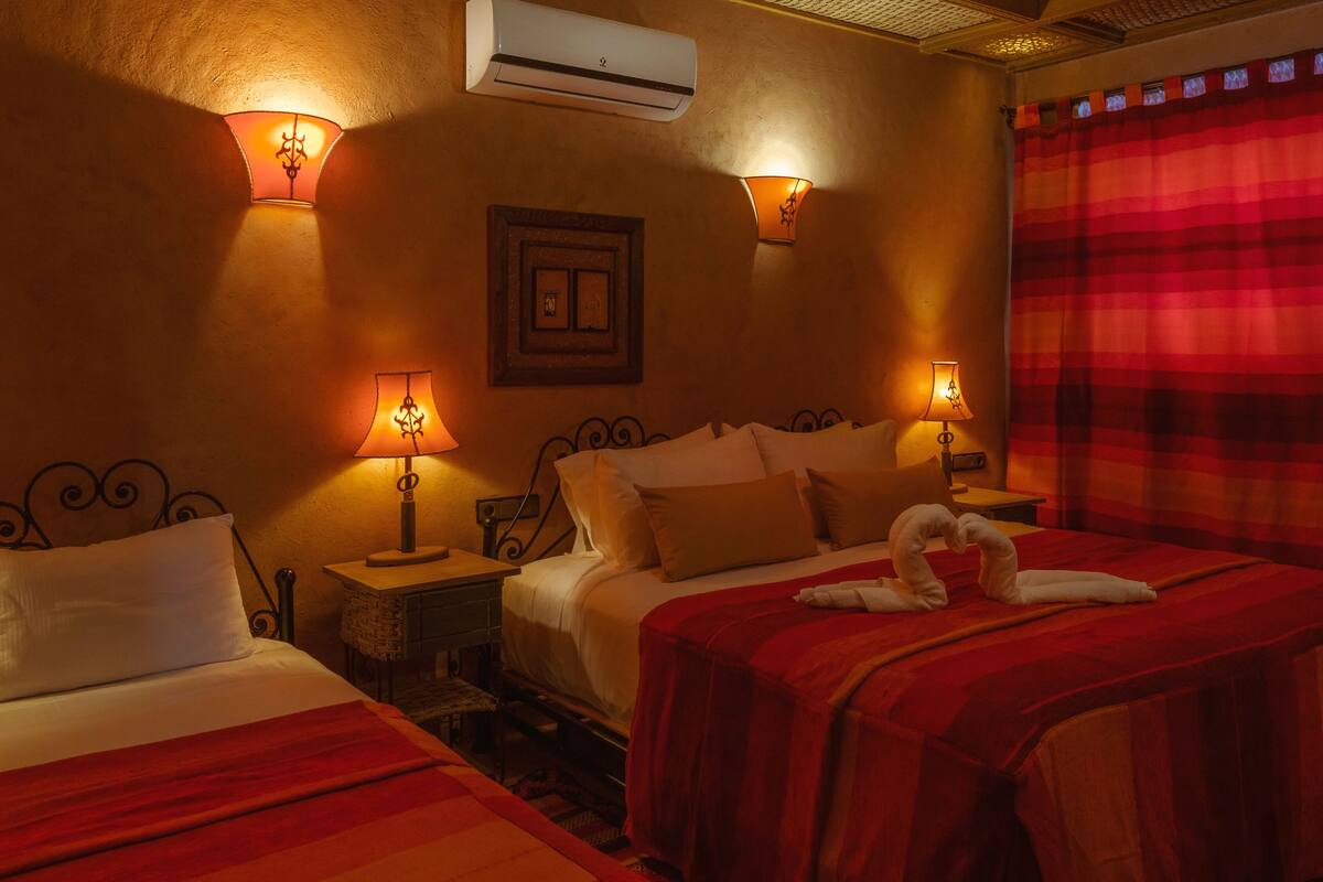 A hotel room with two beds, each with a red blanket, two wall-mounted lamps, and an air conditioning unit.