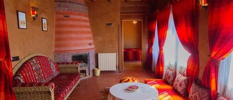 A cozy room with a fireplace, a sofa, a dining table, and a large window with red curtains.