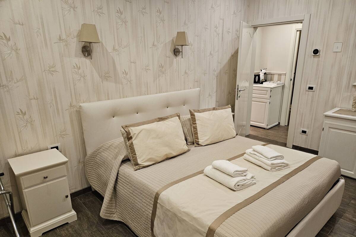 A neatly made bed with a quilted bedspread and pillows, a bedside table, a nightstand, and a bathroom visible through an open door.