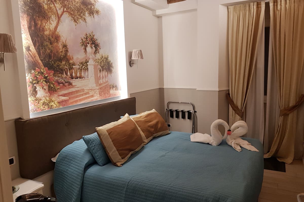 A bedroom with a bed, a headboard, a window with curtains, a wall-mounted radiator, and a painting of a garden scene.
