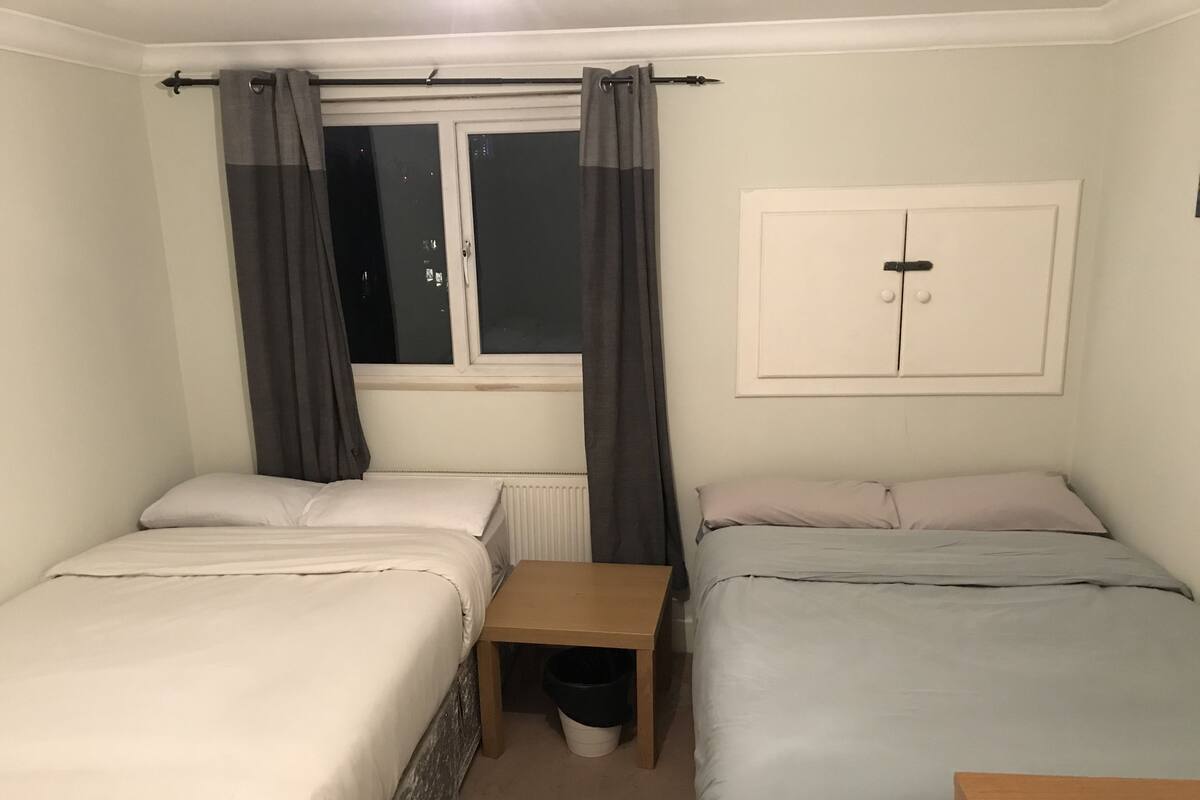 A small, clean room with two single beds, a small table, a radiator, and a window with curtains.