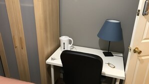 A small, neatly organized room with a white desk, a black office chair, a blue desk lamp, a white electric kettle, and a door with a brass handle. - Rooms at 5 Hammersmith Road - Hostel (London)