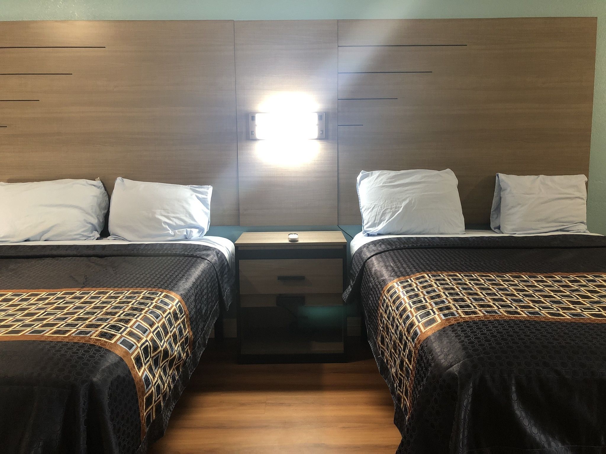 A hotel room with two beds, each with a patterned bedspread and white pillows. There is a nightstand between the beds with a small lamp.