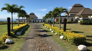 A pathway leading to a building with a modern design, surrounded by palm trees and yellow flowering plants. - Fiji Luxury Pool Villa 2 (Lautoka)