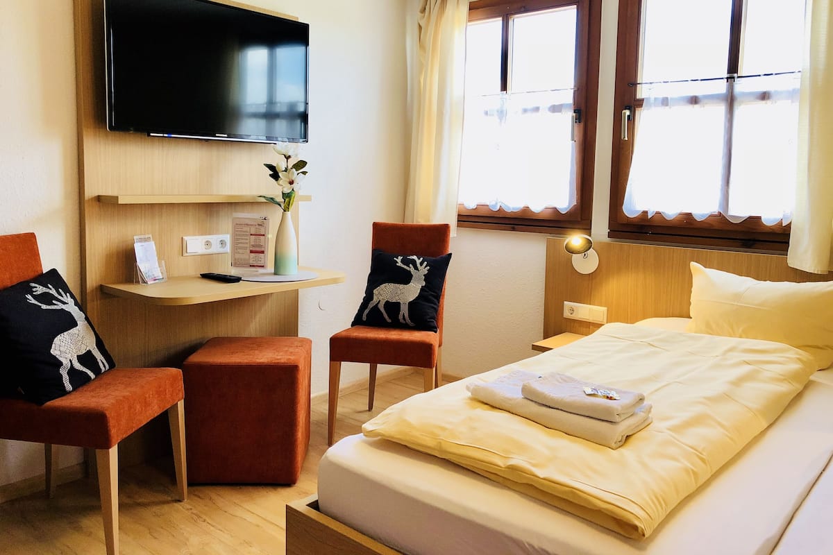 Une chambre d’hôtel avec un lit, deux chaises, un bureau avec une télévision et une fenêtre avec des rideaux.
