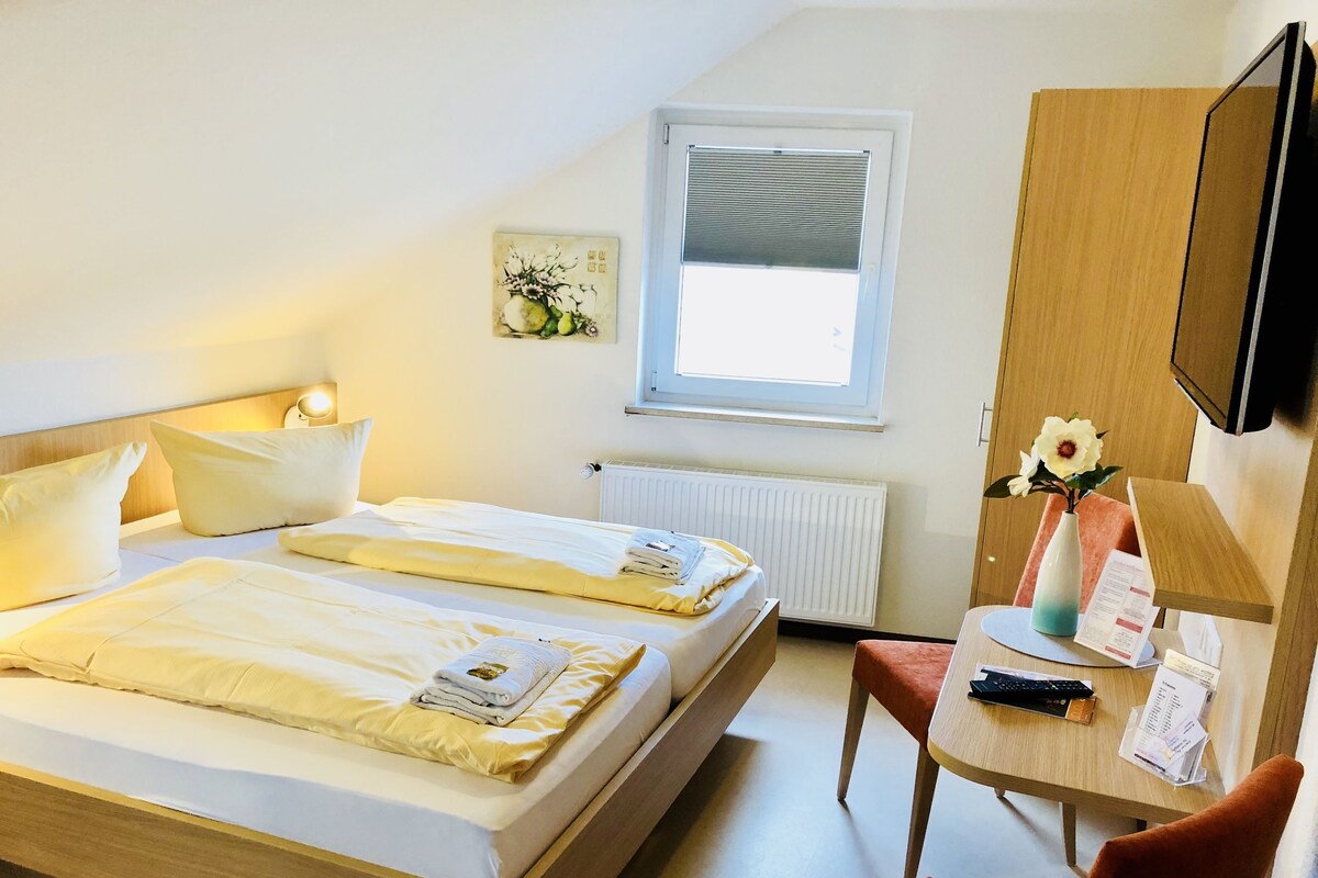 A hotel room with two beds, a desk, a chair, a radiator, a window, and a wall-mounted TV.