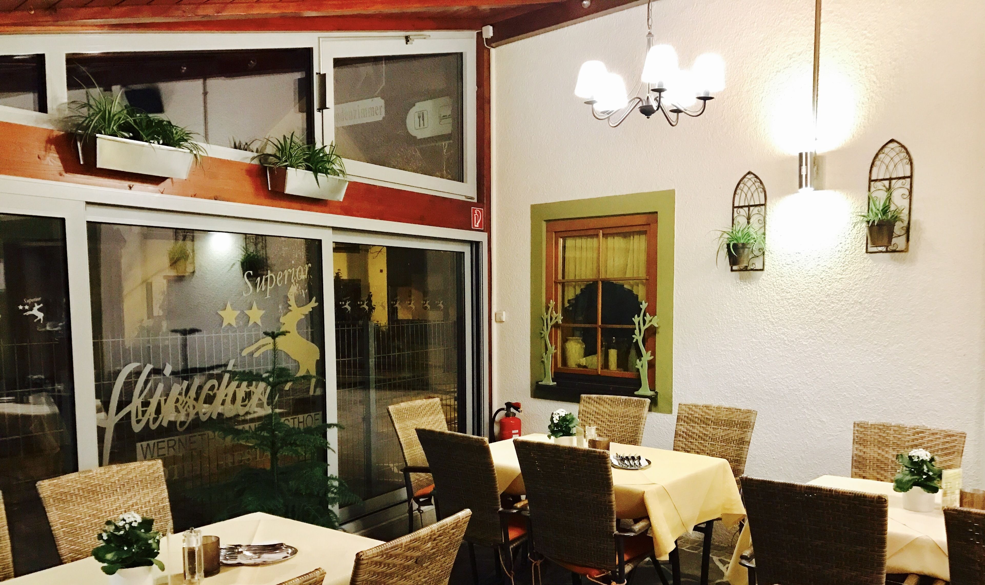 A restaurant interior with wicker chairs, tables with white tablecloths, and potted plants.