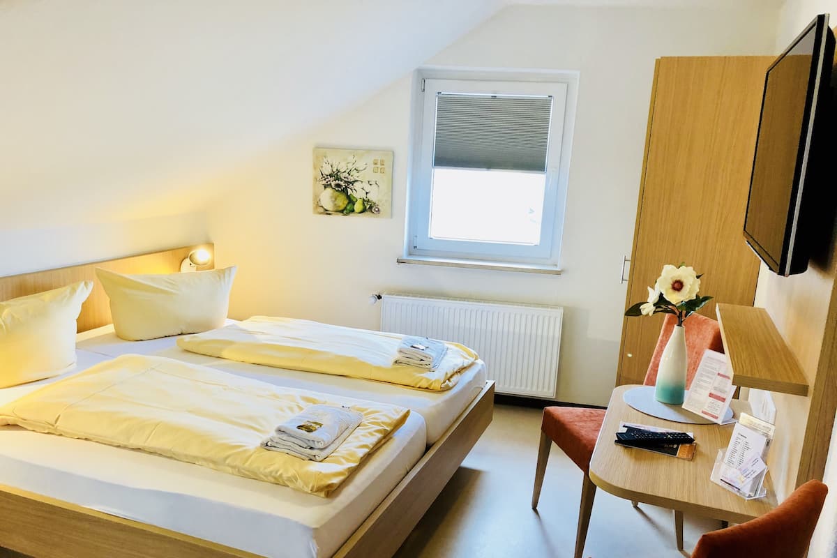 Une chambre d’hôtel avec deux lits, un bureau, une chaise, un radiateur, une fenêtre, un téléviseur fixé au mur et une petite œuvre d’art.