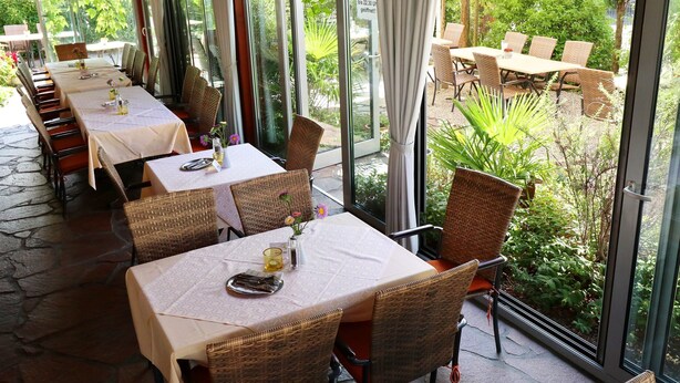 An outdoor dining area with tables and wicker chairs, large windows, and a view of greenery. - Hotel Werneths Landgasthof Hirschen (Rheinhausen)