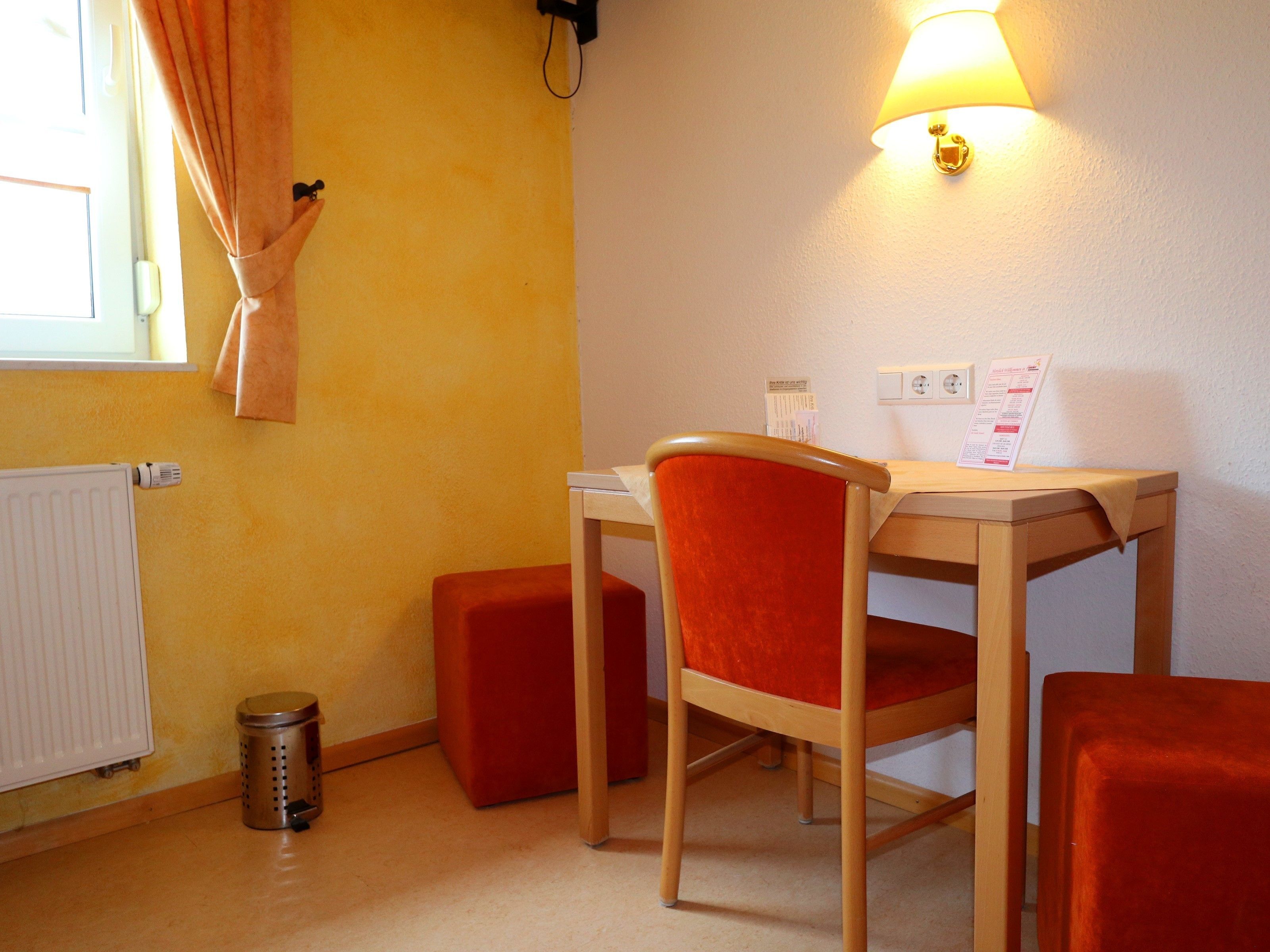Una habitación pequeña con un escritorio de madera, una silla roja, un radiador, un cubo de basura y una lámpara en la pared.