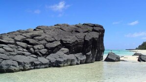 A large rock formation near a clear, shallow beach with a clear sky. - Nikao Luxury Villa (Rarotonga)