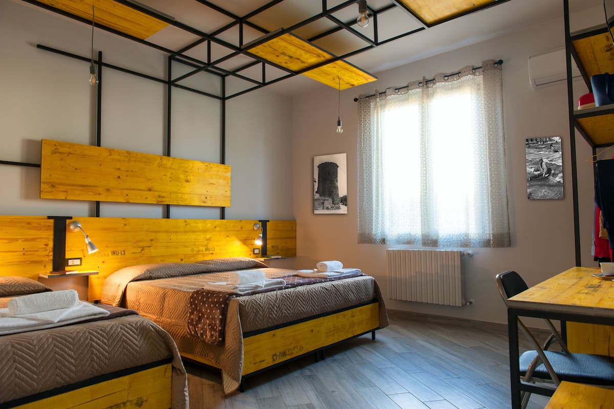 Quarto de hotel com cabeceira de madeira, cama com colcha estampada, mesa de cabeceira com abajur e janela com cortinas.