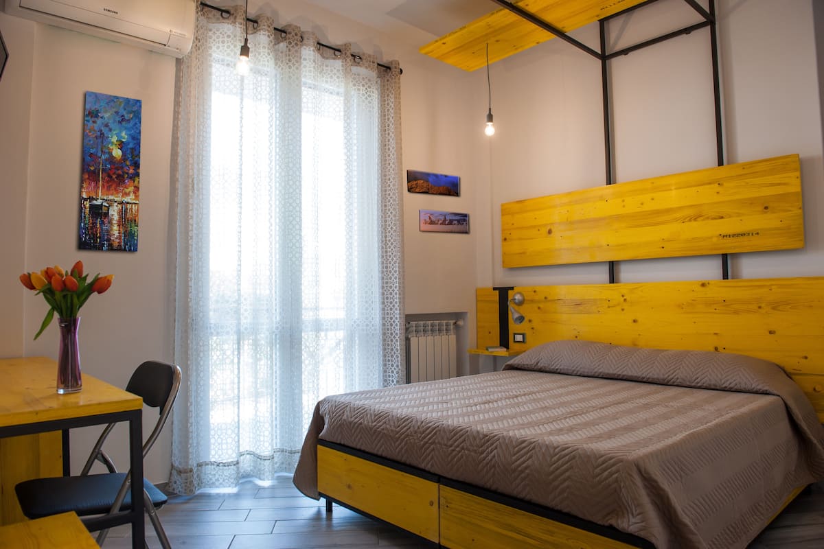 Quarto com cama, escrivaninha e cadeira. Tem janela com cortinas, radiador e um quadro na parede.