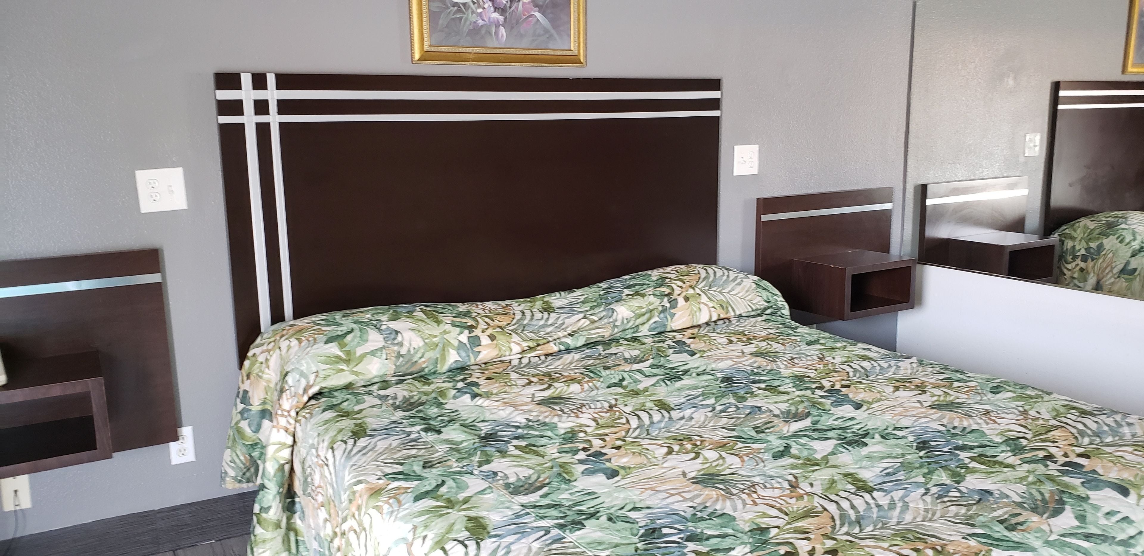 Un lit bien fait, recouvert d’une couvre-lit à motifs floraux, avec une tête de lit sombre agrémentée de touches blanches, et une petite table de chevet dotée d’un miroir.