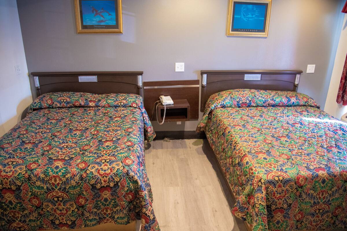 A hotel room with two beds, each with a patterned bedspread, a small bedside table with a phone, and two framed pictures on the wall.
