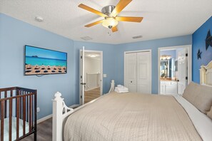 Quarto com cama, berço, ventilador de teto, quadro e porta que dá para outro cômodo.