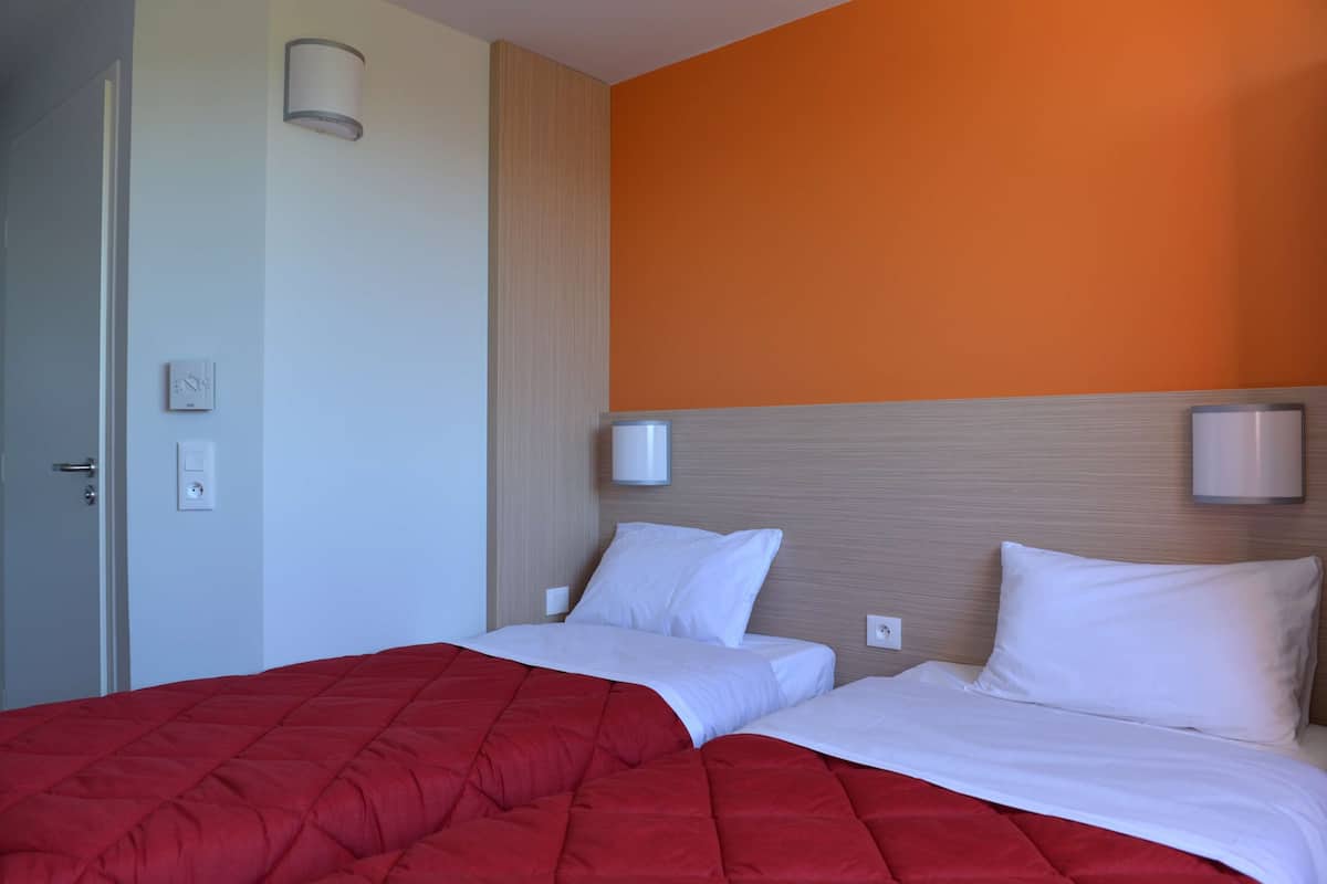 Ein Hotelzimmer mit zwei Betten, einer roten Tagesdecke, weißer Bettwäsche, einer orangefarbenen Akzentwand und einer Tür mit Lichtschalter.