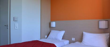 A hotel room with two beds, a red bedspread, white bedding, an orange accent wall, and a door with a light switch.