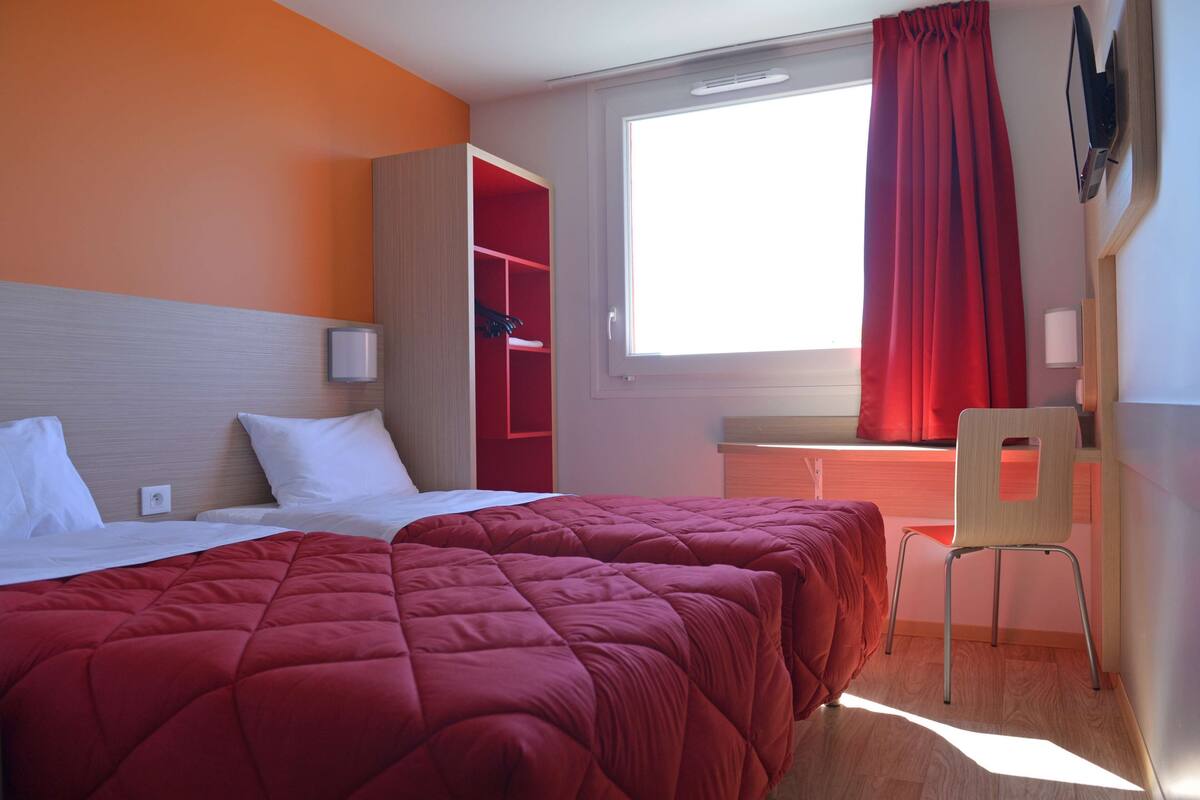 Ein Hotelzimmer mit zwei Betten, einem Schreibtisch, einem Stuhl und einem Vorhangfenster.