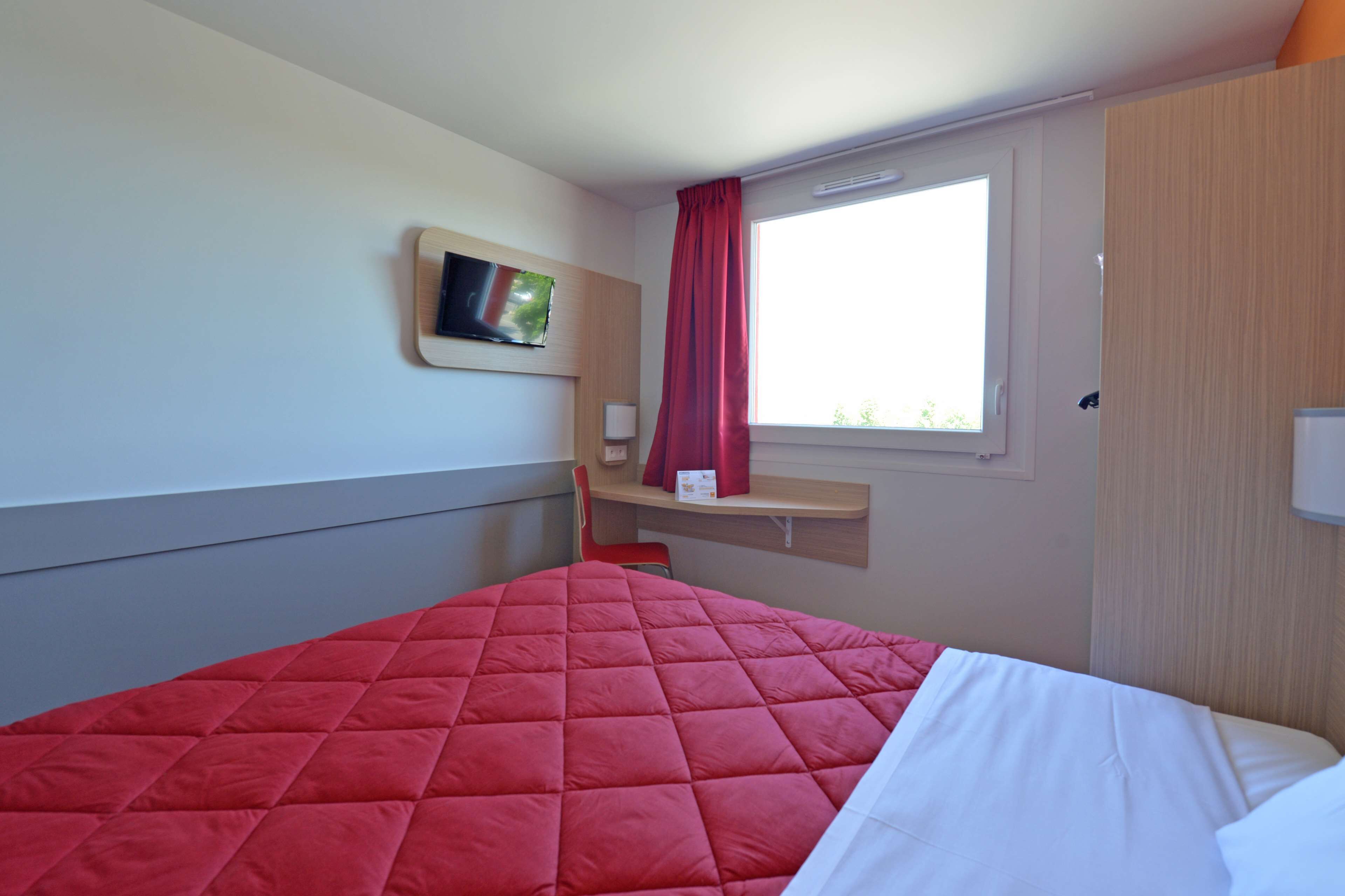 Een hotelkamer met een bed, een aan de muur bevestigde televisie, een raam met rood gordijn en een kleine tafel met een lamp.