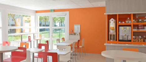 Un espace intérieur lumineux, avec des murs orange, des tables blanches et des chaises rouges. Une grande fenêtre offre une vue sur la verdure à l’extérieur.
