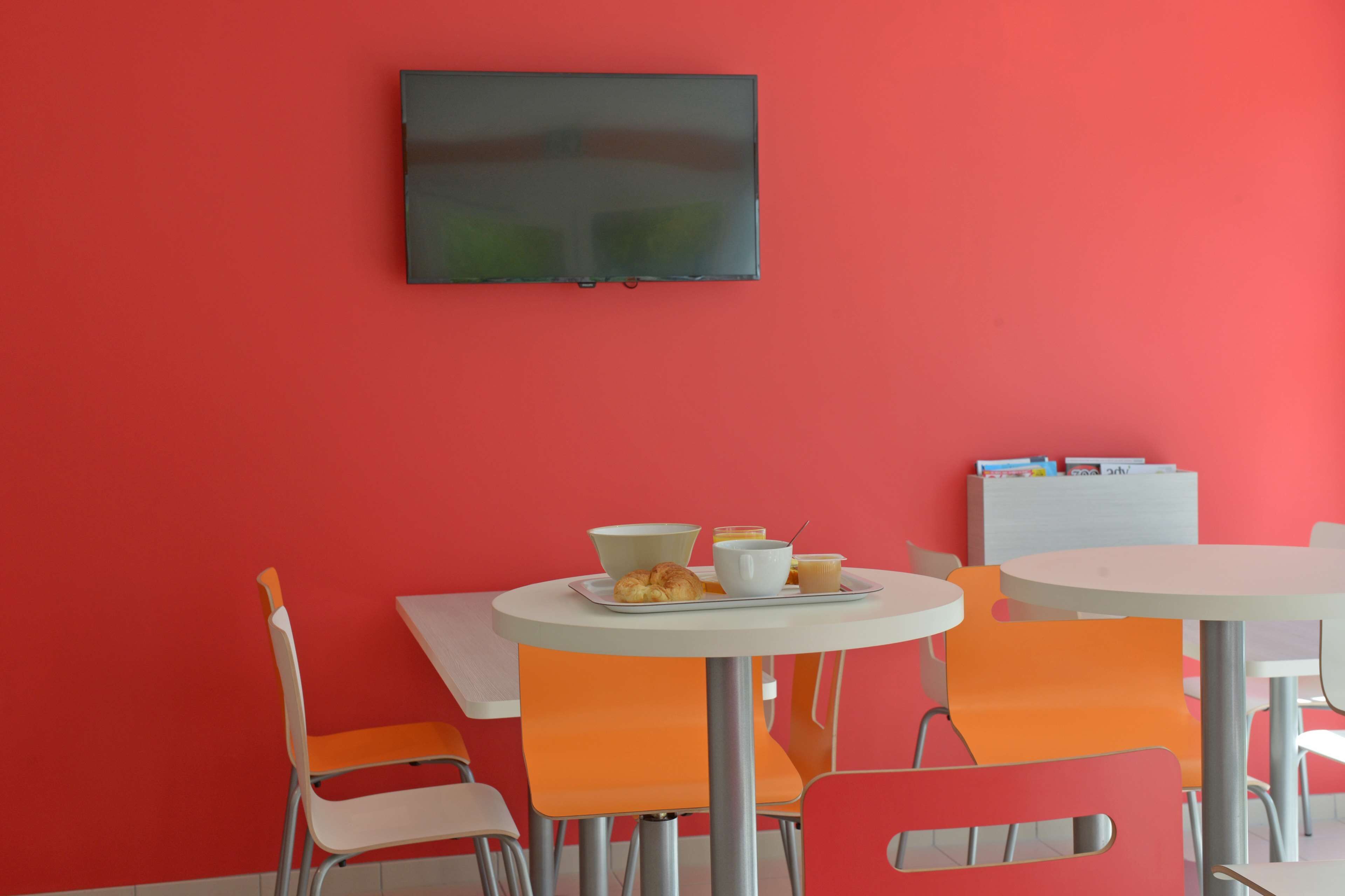 A dining area with a red wall, white and orange chairs, a round table with breakfast items, and a flat-screen TV mounted on the wall.