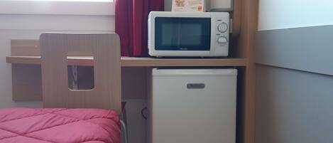 A compact room with a bed, a small white refrigerator, a microwave, and a wooden chair.